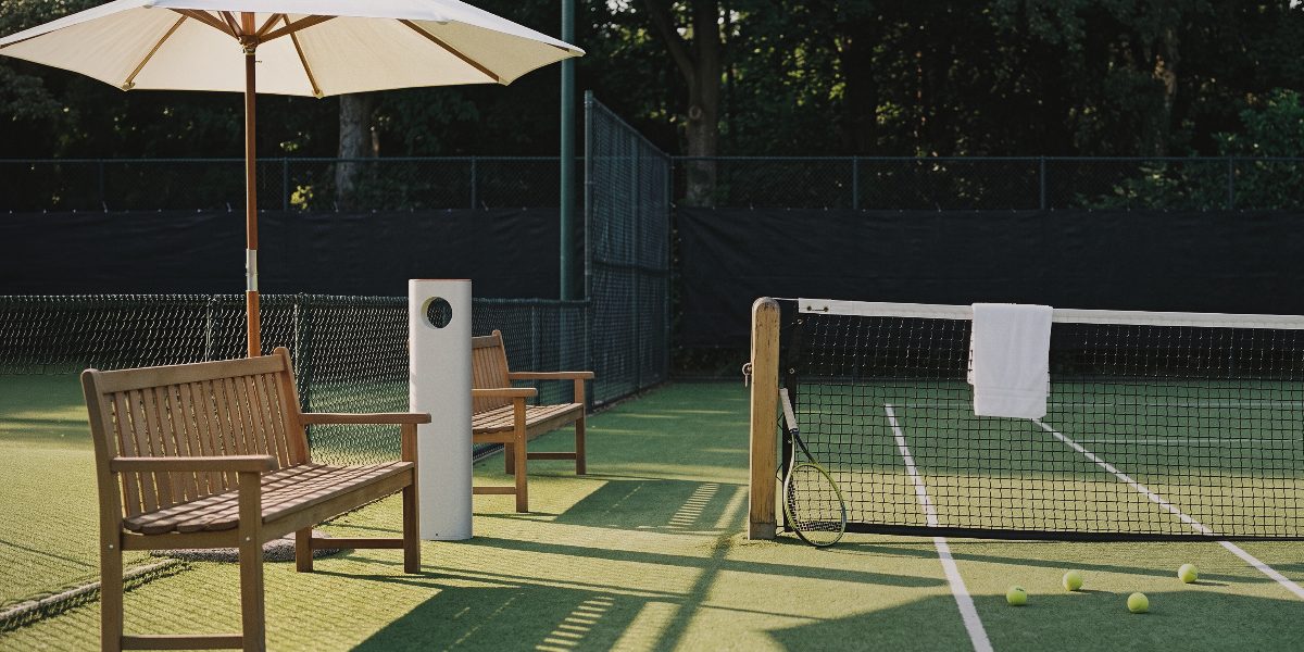 A dispenser courtside beside a bench and parasol on a tennis court